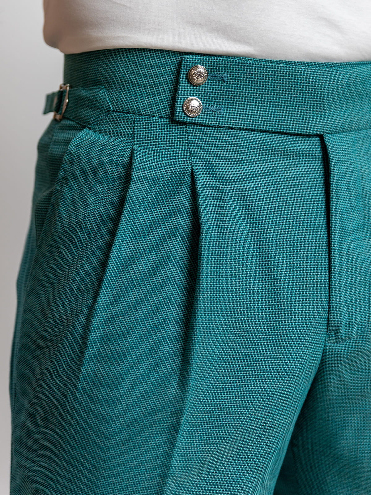 Teal pleated pants with a white shirt underneath on a light gray background